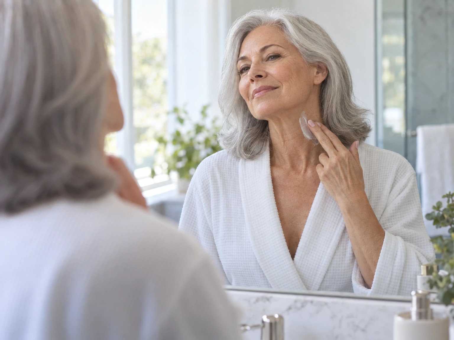 A woman in her sixties smoothing the cream up the side of her neck at a bathroom mirror